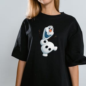 Peut inclure: Un t-shirt noir &agrave; manches courtes avec une impression graphique du bonhomme de neige blanc Olaf du film La Reine des Neiges. Le personnage a un nez en carotte, des bras en brindilles et trois boutons noirs sur le corps, centr&eacute; sur le devant.
