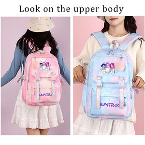 May include: A backpack featuring a gradient color scheme with a graphic of three figures and the text "HUNTRAX". The bag includes adjustable rainbow-striped straps, a front pocket with buckle closures, and a fluffy pom-pom charm attached to the zipper.