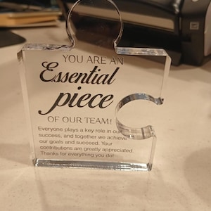 May include: A clear acrylic puzzle piece award featuring black printed text that reads: YOU ARE AN Essential piece OF OUR TEAM! Everyone plays a key role in our success, and together we achieve our goals and succeed. Your contributions are greatly appreciated.