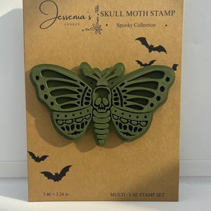 May include: An olive green skull moth stamp on a tan backing card. The card features the text Jessenia's Corner, SKULL MOTH STAMP, Spooky Collection, and 5.46 x 3.24 in. The moth design has a skull on its thorax and intricate wing cutouts.