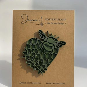 May include: A dark olive green pottery stamp featuring a heart-shaped honeycomb pattern with a bee and flowers. The stamp is mounted on a tan card labeled Jessenia's Corner Pottery Stamp Bee Garden Design, measuring approximately 4.13 inches.