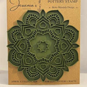 May include: An olive green mandala pottery stamp on a tan cardboard backing. The text reads Jessenia's Corner, POTTERY STAMP, Boho Mandala Design, APROX. 6.3 X 6.3 INCHES, and FOR CLAY, POTTERY, CRAFTS. The stamp features a symmetrical floral pattern.