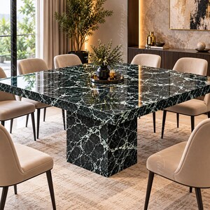 Op de afbeelding: Een vierkante eettafel met een gepolijst donkergroen marmeren oppervlak met opvallende witte aders, ondersteund door een bijpassende vierkante voet. De tafel is omringd door zes gestoffeerde stoelen met lichtbeige bekleding en donkere poten.