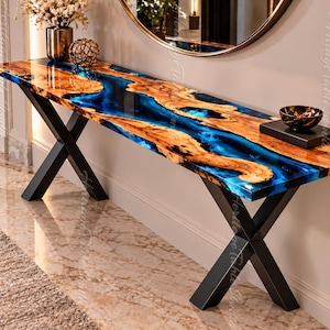 Puede incluir: Una mesa consola con un tablero de madera de borde natural y una incrustación de resina azul vibrante, sostenida por dos patas de metal negro en forma de X. La superficie es pulida y brillante, mostrando vetas de madera y resina azul.