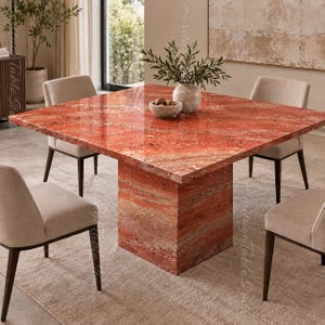 Op de afbeelding: Een vierkante eettafel met een gepolijst oppervlak van rode travertijnsteen en een bijpassende voet. Vier gestoffeerde beige stoelen met donkerhouten poten staan rondom de tafel, waarop een kleine kom en een plant zijn geplaatst.