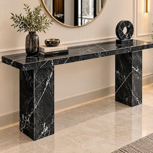 May include: A rectangular console table crafted from black marble with prominent white veining. The table features a long, flat top supported by two thick, block-style legs. On the surface sits a dark glass vase with greenery, a small bowl, two stacked books, and a circular black marble sculpture.