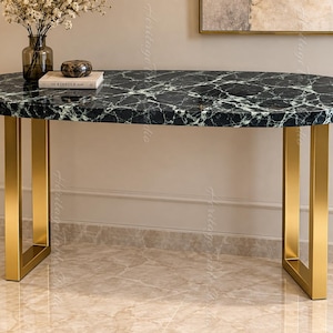 May include: An oval console table featuring a black marble top with white veining and two U-shaped gold metal legs. The table surface holds a glass vase with dried flowers and a book titled The Kinfolk Table with a small dark stone object.