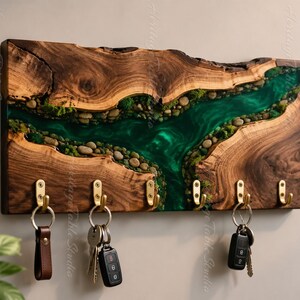 Könnte beinhalten: Ein rechteckiger Wand-Schlüsselhalter aus einer natürlichen Holzplatte mit Baumkante. Eine mittige, flussartige Einlage aus durchscheinendem grünem Harz enthält kleine graue Flusskiesel und grünes Moos. Sechs Messinghaken halten verschiedene Schlüssel.