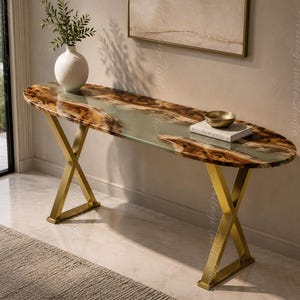 Può includere: Un tavolo console ovale con piano in legno a bordo naturale e inserto centrale in resina traslucida, sostenuto da due gambe in metallo dorato a forma di X. Un vaso bianco con vegetazione e un libro con una ciotola poggiano sul piano.
