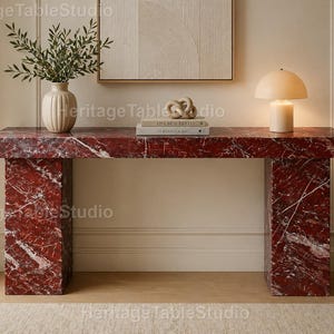 May include: A rectangular console table crafted from deep red marble with prominent white veining. The table features a thick, flat top supported by two wide, solid rectangular legs, creating a sturdy and substantial furniture piece.