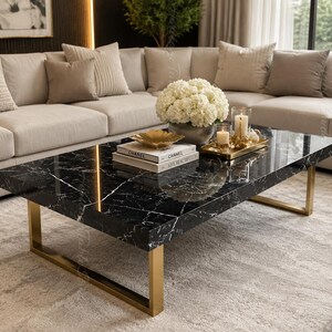 May include: A rectangular coffee table featuring a black marble top with white veining and a gold-toned metal frame. The table surface holds two stacked books labelled Chanel, a gold bowl, a vase of white flowers, and a gold tray with candles.