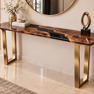Può includere: Un tavolo consolle rettangolare con piano in legno a bordo naturale e inserto centrale in resina nera, sostenuto da due gambe in metallo dorato lucido. Sulla superficie sono presenti un vaso dorato con fiori bianchi e due libri.