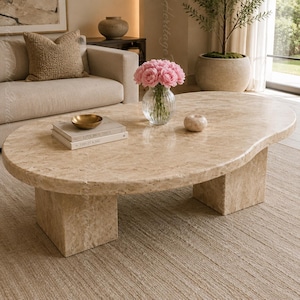 May include: An oval coffee table crafted from beige travertine stone with a textured, porous surface. The table features a thick, rounded tabletop supported by two substantial, rectangular block legs made of the same matching stone material.