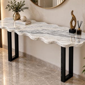May include: A console table featuring a white marble top with grey veining and a scalloped edge design. The table is supported by two black metal U-shaped legs. On the surface sits a gold vase with greenery, a book, a sphere, and decor.