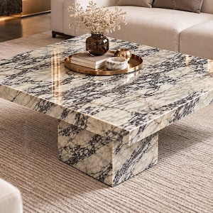 May include: A square coffee table made of polished white marble with prominent black and grey veining. The table features a thick, flat top supported by a central, block-shaped pedestal base, creating a low-profile, solid structure.