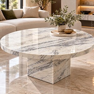 May include: A round coffee table featuring a polished white marble surface with prominent grey veining. The table is supported by a thick, square-shaped pedestal base made of the same matching marble material, creating a cohesive and solid structure.