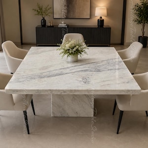 Op de afbeelding: Een vierkante eettafel met een wit marmeren blad met grijze aders en een bijpassende voet, omringd door vier crèmekleurige gestoffeerde stoelen met zwarte poten. In het midden staat een kleine witte vaas met groen blad.