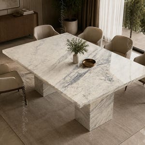 Pode incluir: Uma grande mesa de jantar retangular com um tampo de mármore branco polido com veios cinzentos e uma base de pedestal de mármore a condizer. Seis cadeirões estofados em bege com pernas de madeira escura estão dispostos à volta da mesa. Um pequeno vaso com vegetação e uma taça de bronze rasa estão colocados sobre a superfície.