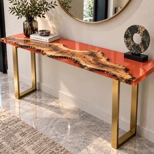 Puede incluir: Una mesa consola rectangular con una superficie de resina naranja vibrante y una losa de madera natural con borde vivo en el centro. La mesa está sostenida por dos patas de metal dorado cepillado en forma de U y tiene un acabado liso y brillante.