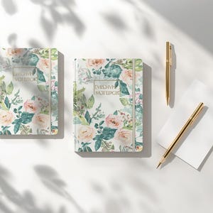 Peut inclure: Deux carnets à couverture rigide ornés d'un motif floral aquarelle dans des tons pêche, vert et blanc, avec le texte "EMBRACE NOTEBOOK" dans un cadre rectangulaire doré. Deux stylos dorés sont posés à côté d'une carte rectangulaire blanche.