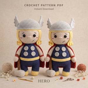 Peut inclure: Deux poupées au crochet avec des cheveux blonds, des casques ailés gris, des tenues bleues avec des accents circulaires argentés, des ceintures jaunes et des capes rouges. Chaque poupée tient un marteau gris. Texte : CROCHET PATTERN PDF, HERO.