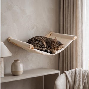 May include: A wall-mounted cat hammock featuring a light beige fabric sling suspended between two smooth, cylindrical wooden dowels. A brown and black tabby cat is curled up asleep in the center of the hammock, which is attached to the wall.