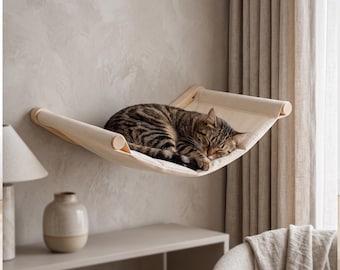 May include: A wall-mounted cat hammock featuring a light beige fabric sling suspended between two smooth, cylindrical wooden dowels. A brown and black tabby cat is curled up asleep in the center of the hammock, which is attached to the wall.