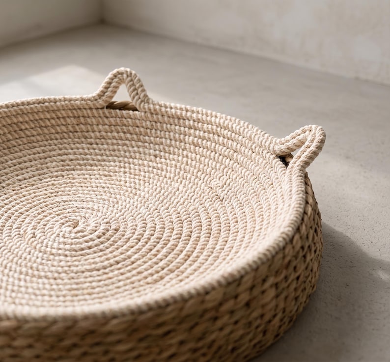 Pode incluir: Uma bandeja redonda e rasa constru&iacute;da com corda bege clara enrolada. A base interna apresenta um padr&atilde;o de corda circular apertado, enquanto os lados externos s&atilde;o feitos de uma fibra tecida de cor natural. Duas pequenas al&ccedil;as triangulares de corda estendem-se para cima a partir da borda.