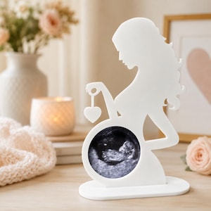 May include: A white silhouette figure of a pregnant person standing on an oval base. The figure features a circular cutout over the abdomen area containing a black and white ultrasound image of a fetus, and a small heart shape hanging from one hand.