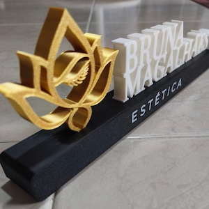 May include: A 3D-printed sign featuring a gold lotus flower icon on the left and white raised lettering that reads BRUNA MAGALHAES. Below the name, the word ESTETICA is printed in white on a long, rectangular black base stand.