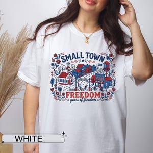 May include: A white short-sleeve t-shirt featuring a graphic illustration of a small town with red, white, and blue buildings, a water tower, and floral accents. The text reads SMALL TOWN FREEDOM and 1776-2026 years of freedom.