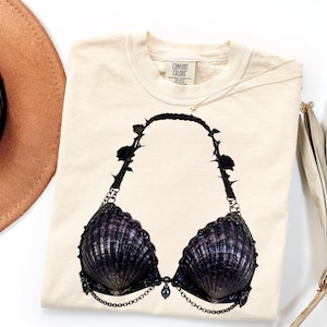 May include: A cream-colored t-shirt featuring a graphic print of a dark purple seashell bikini top. The bikini design includes black rose and thorn accents on the straps, metallic chain detailing, and a central dark bead hanging from the center.