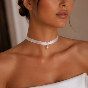 Può includere: Una collana girocollo in nastro di raso bianco con un ciondolo di perla a forma di goccia sospeso da un piccolo connettore in metallo argentato. Il nastro liscio e sottile aderisce al collo, mettendo in risalto la perla bianca lucente.