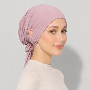 May include: A dusty rose head covering made of soft, stretchable fabric. The head wrap features a fitted crown that contours to the head and includes a gathered section at the nape of the neck with two long, thin fabric ties that hang down.