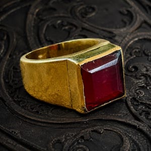 May include: A gold ring featuring a large, square-cut, deep red gemstone set into a thick, polished band. The ring has a substantial, solid construction with a smooth, reflective surface that highlights the vibrant color of the central stone.