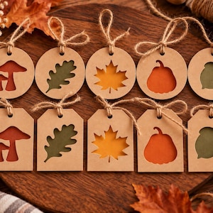 Può includere: Dieci etichette regalo in carta kraft marrone di forma rotonda e rettangolare, ognuna con un motivo autunnale fustellato che include funghi, foglie di quercia, foglie d'acero, zucche e ghiande, tutte legate con cordini di juta naturale.