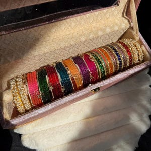 May include: A collection of multi-coloured bangles arranged in a row inside a rectangular, velvet-lined storage box. The bangles feature various vibrant hues including red, green, orange, pink, and blue, with some adorned with small white pearls.