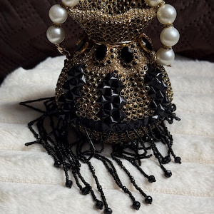 May include: A decorative pouch bag featuring a gold-tone metal frame encrusted with small black crystals and large black faceted studs. The handle is made of large white faux pearls. The base is adorned with long, dangling black beaded fringe.