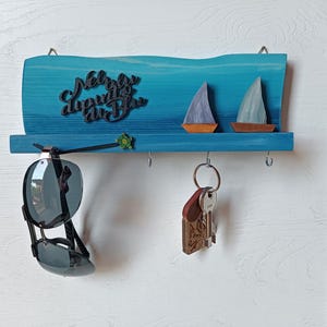 Op de afbeelding: Een blauwe houten wand-sleutelhouder met een golvende bovenrand, voorzien van twee kleine houten zeilbootjes, een schildpadaccent en de tekst "Nel blu dipinto di blu". Het heeft vier metalen haken, met een zonnebril en een sleutelbos eraan.