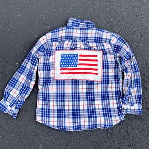 May include: A long-sleeve button-down shirt featuring a blue, white, and red plaid pattern. A rectangular patch is sewn onto the back, displaying a graphic of the American flag with red stripes and white stars on a blue field, with the text TEXTILES.