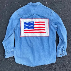 May include: A light blue denim long-sleeve button-down shirt featuring a rectangular white patch on the back. The patch displays a stylized American flag with blue stars and red stripes, with the word TEXTISLES printed in small black letters.