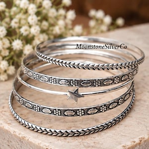 May include: A set of five silver bangles featuring varied textures including braided patterns, floral motifs, and a central star charm. The text MoonstoneSilverCo is visible on the top bangle. Each bangle has an oxidised finish to highlight the intricate details.