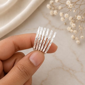 May include: A set of six thin silver rings, each featuring a textured, hammered surface with small indentations along the outer band. The rings are arranged in a parallel row, showcasing a bright, reflective metallic finish and a consistent design.