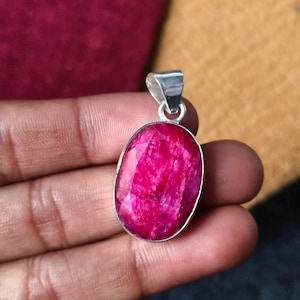 May include: An oval-shaped pendant featuring a faceted, vibrant magenta-pink gemstone set in a polished silver-toned metal frame. The pendant includes a simple silver-toned bail at the top for attachment to a chain or cord, showcasing a smooth finish.