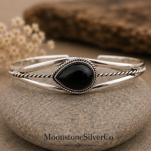 Può includere: Un bracciale a fascia in argento con una pietra nera centrale a forma di goccia incastonata in un bordo a corda intrecciata. Il cinturino è composto da due fili d'argento lisci che affiancano un filo intrecciato. Il testo MoonstoneSilverCo è stampato in basso.