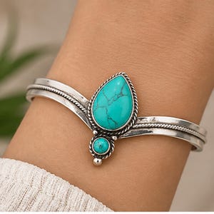 May include: A silver cuff bracelet featuring a large teardrop-shaped turquoise stone and a smaller round turquoise stone. The metal band has a twisted rope texture and a polished finish, with the stones set in a decorative braided silver frame.