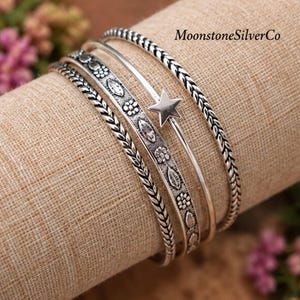 以下が含まれることがあります： 4本のシルバー調バングルセット。編み込み模様のバンド2本、花と葉の模様が施されたバンド1本、小さな五芒星があしらわれた滑らかなバンド1本で構成されています。MoonstoneSilverCoというテキストが表示されています。