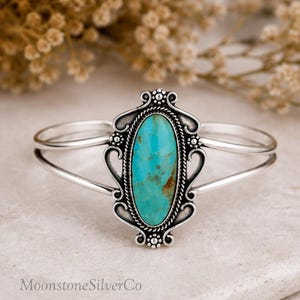 May include: A silver cuff bracelet featuring a central oval turquoise stone with brown matrix inclusions. The stone is framed by a twisted rope border and decorative silver scrollwork with small bead accents on a polished silver band.