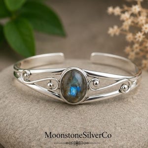 May include: A silver cuff bracelet featuring a central oval labradorite gemstone with blue iridescence. The band has a polished finish and decorative scrollwork details on both sides of the stone. The text MoonstoneSilverCo is visible.
