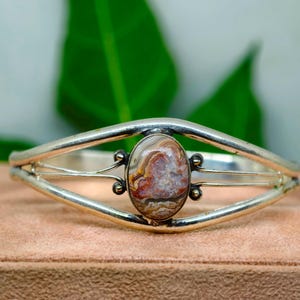 Puede incluir: Una pulsera de brazalete de plata con una piedra central ovalada que presenta patrones intrincados y arremolinados en tonos marrón, crema y naranja rojizo. La banda de metal se divide en dos brazos pulidos que rodean la piedra.